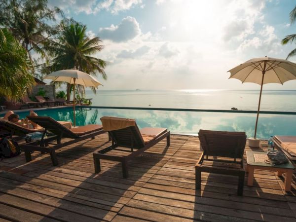 A tranquil poolside scene with lounge chairs, umbrellas, and a view of the ocean, set against a sunny sky with scattered clouds.