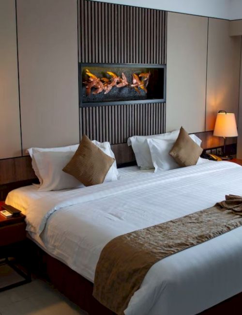 A neatly arranged hotel room with a king-size bed, decorative pillows, lamps on side tables, wall art, and draped curtains by a window.