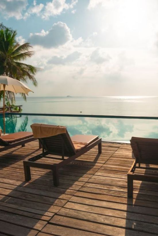 A serene poolside setting with lounge chairs, umbrellas, and a view of the ocean under a partly cloudy sky at sunrise or sunset.