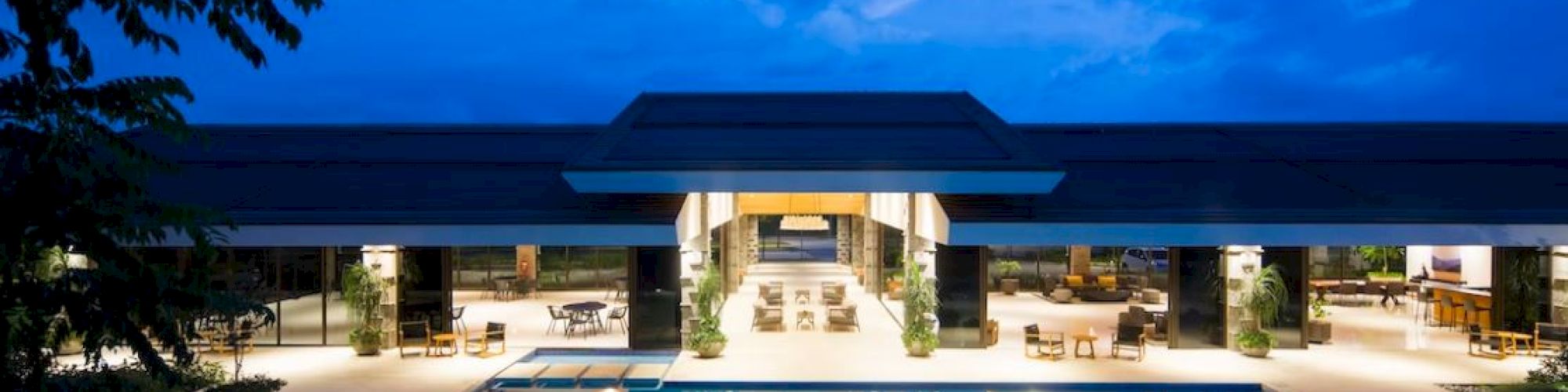 The image shows a luxurious building with a large swimming pool, surrounded by lounge chairs, illuminated at night under a blue sky.