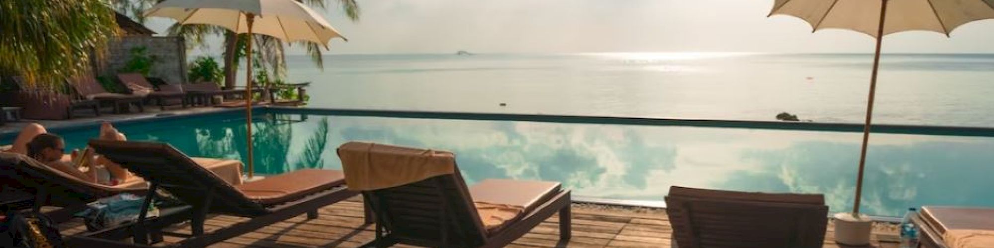 Poolside scene with lounge chairs, umbrellas, and palm trees overlooking a calm ocean during daylight.
