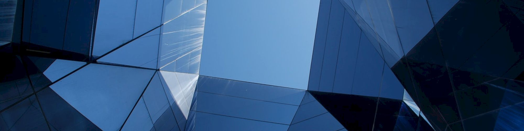 The image shows a geometric architectural structure with reflective glass panels, forming a tunnel-like view towards the sky.