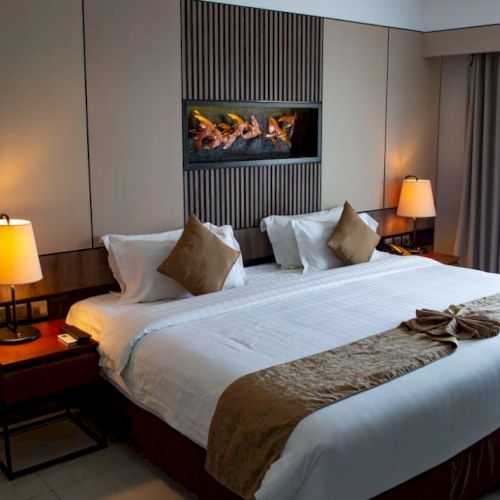 A neatly made bed in a cozy hotel room with warm lighting, pillows, and a wood-accented headboard with a decorative art piece above it.