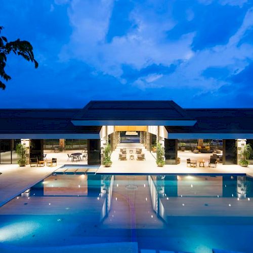 A modern building with a central open area is reflected in a large, illuminated outdoor pool during twilight.