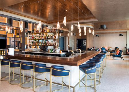 The image shows a modern bar with a central counter, blue bar stools, hanging lights, and shelves stocked with bottles.