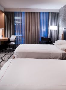 The image shows a modern hotel room with two beds, a flat-screen TV, desk, and large window with curtains, creating a cozy atmosphere.