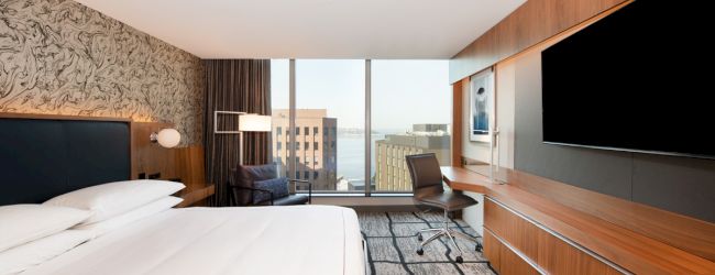 A modern hotel room with a large bed, flat-screen TV, desk, and city view from a window, featuring wood furnishings and patterned carpet.