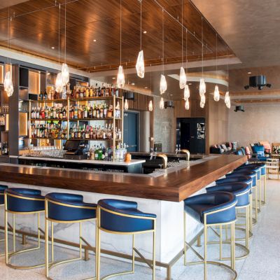A modern bar with a stylish design, featuring a central counter, shelves with bottles, pendant lights, and seating area.