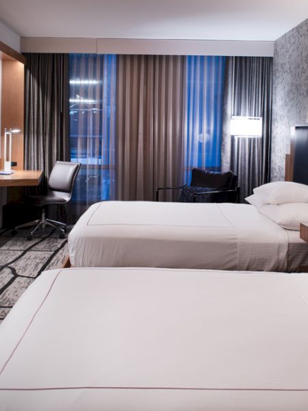 The image shows a modern hotel room with two beds, a wall-mounted TV, a desk, and large curtains, all in a stylish, contemporary design.