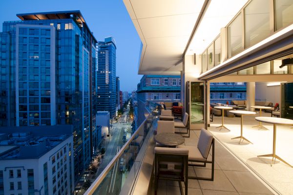 A modern rooftop terrace with seating, overlooking a cityscape of tall buildings at dusk.
