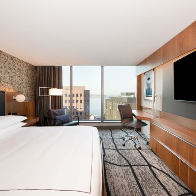 The image shows a modern hotel room with a large bed, a desk, a big TV, and a window overlooking city buildings.