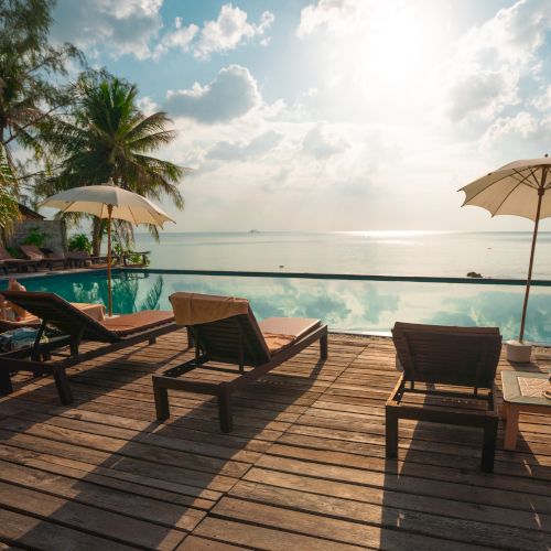 A serene beach scene with lounge chairs, umbrellas, and palm trees overlooking a tranquil ocean under a bright sky with fluffy clouds.