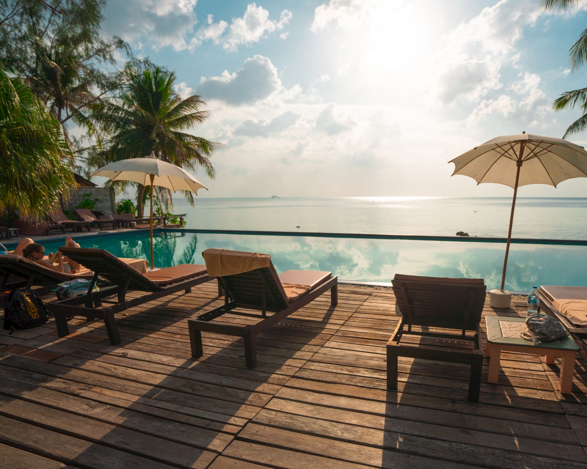 A serene beachside scene with lounge chairs, umbrellas, a pool, palm trees, and an ocean view under a partly cloudy sky.