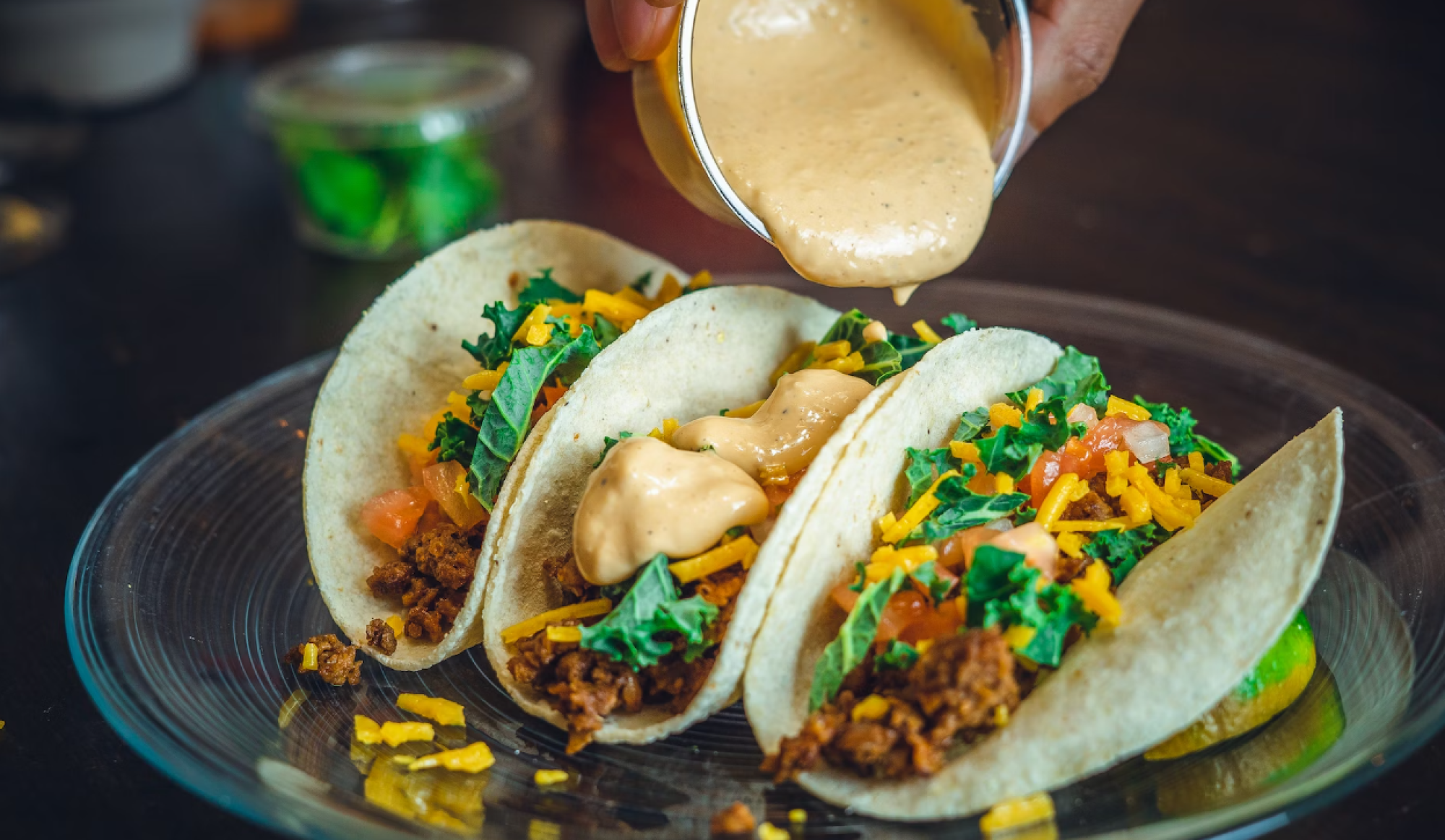 Three tacos filled with meat, cheese, and greens, with sauce being poured over them from a cup.