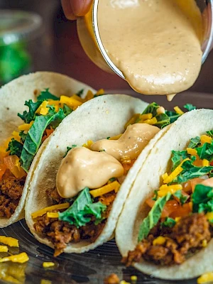 Three tacos filled with ground meat, cheese, and greens are on a plate, with a sauce being poured over them from a small cup.