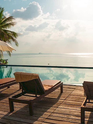 A serene beachside pool setting with lounge chairs, umbrellas, and palm trees, overlooking a calm sea under a partly cloudy sky.