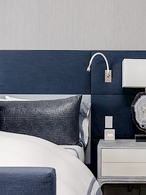 A stylish bedroom features a bed with navy headboard, pillows, a modern side table with a lamp, and a mounted reading light.