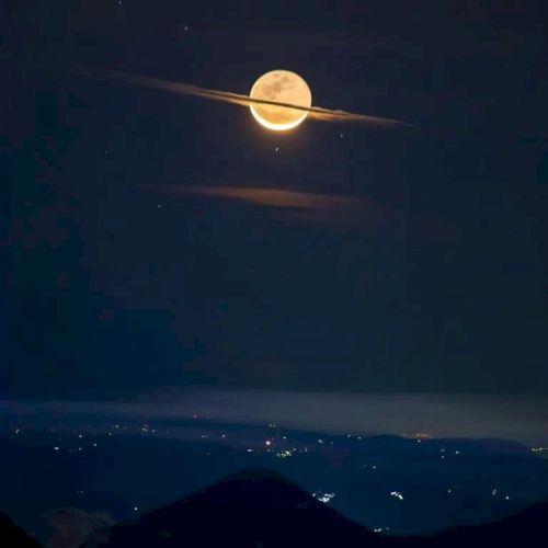 The image shows a full moon with clouds forming an illusion of Saturn's rings, above a dark landscape with distant city lights.