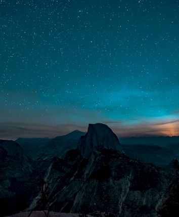 A starry night sky over mountains, with a glowing horizon and silhouetted trees in the foreground.