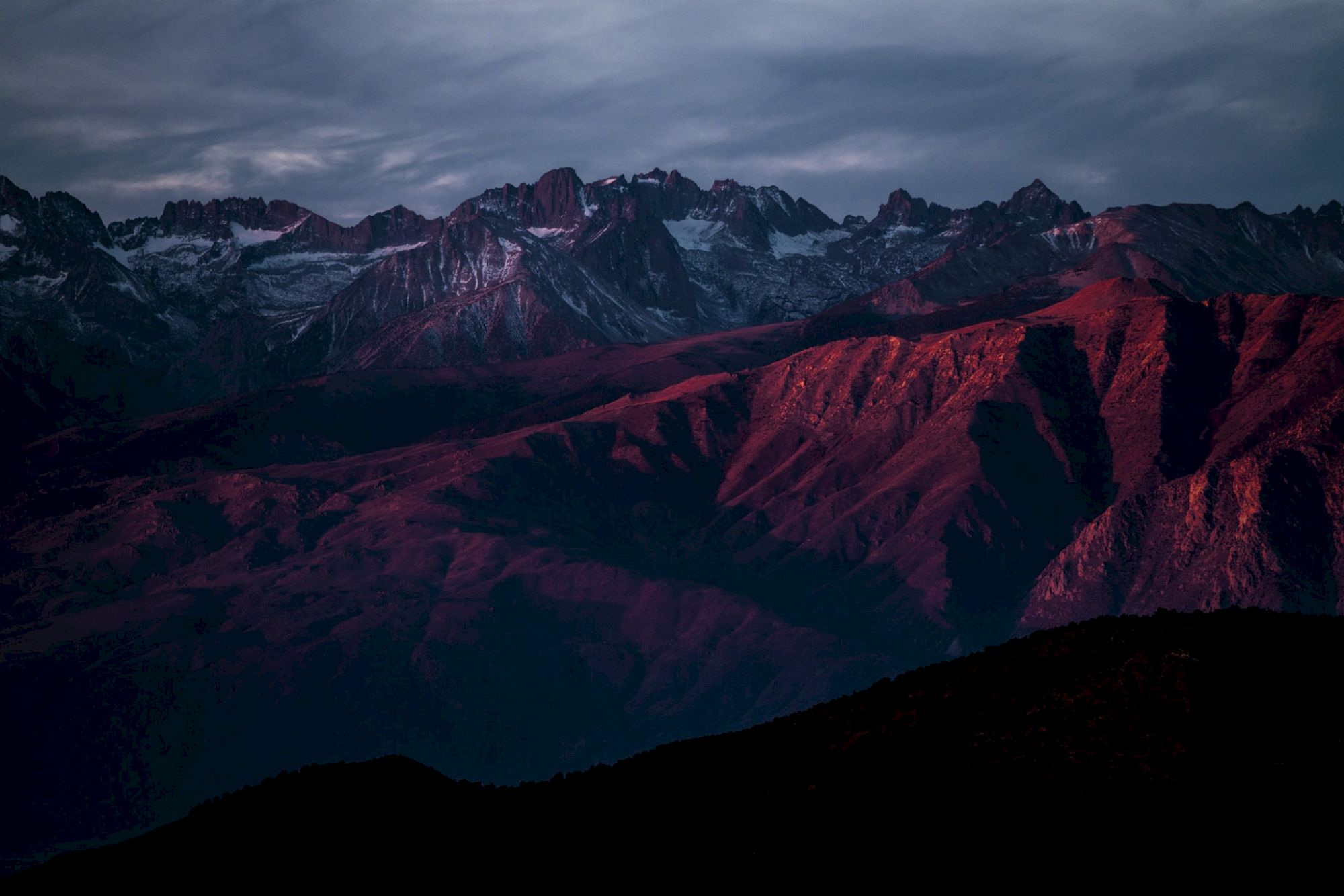 A mountain range bathed in a reddish sunset glow beneath a cloudy sky.
