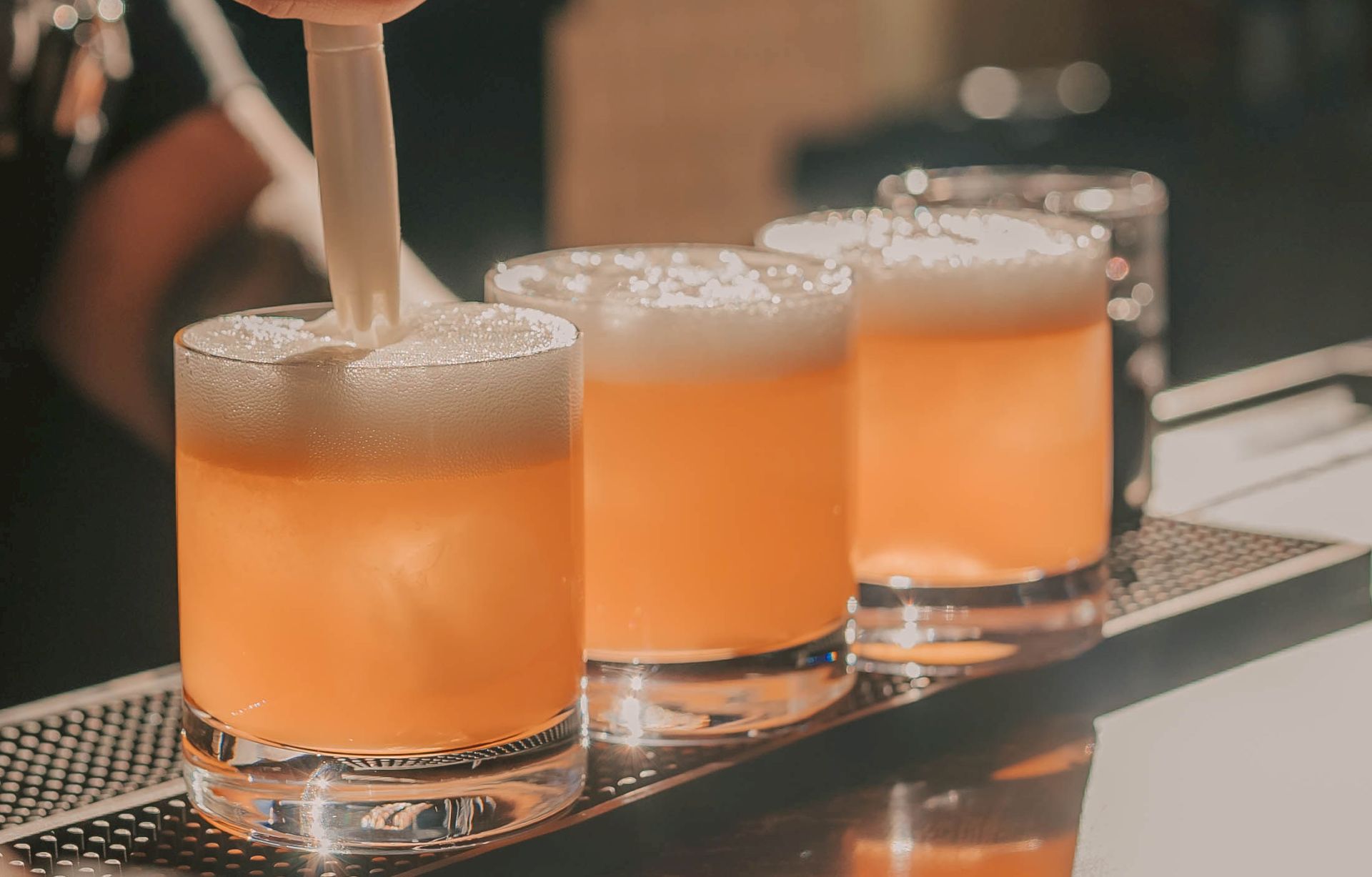 A bartender pours a foamy amber-orange cocktail into a glass on a bar, with another drink behind it.