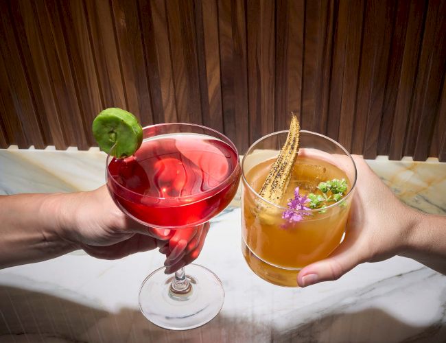 Two hands toasting with cocktails: one red drink with a lime and one amber drink with garnishes, on a marble surface.