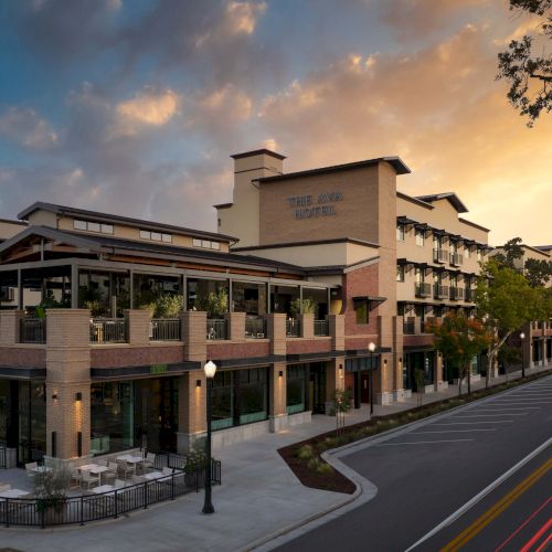 A modern, multi-story shopping and dining complex with brick accents sits beside a tree-lined street, basking in a warm sunset.