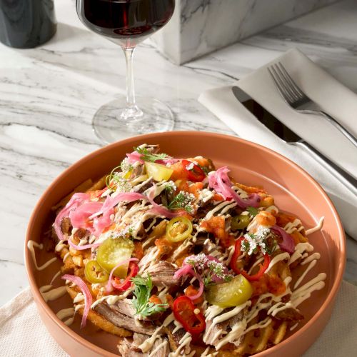 A dish with fries topped with various vegetables and sauce, alongside a glass of red wine on a marble table.