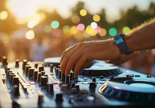 A DJ adjusts controls on a mixer at an outdoor event, with colorful lights and a blurred crowd in the background.