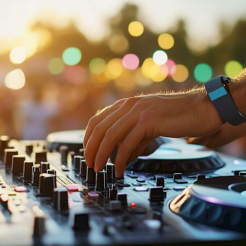 A DJ adjusts controls on a mixer at an outdoor event, with colorful lights and a blurred crowd in the background.