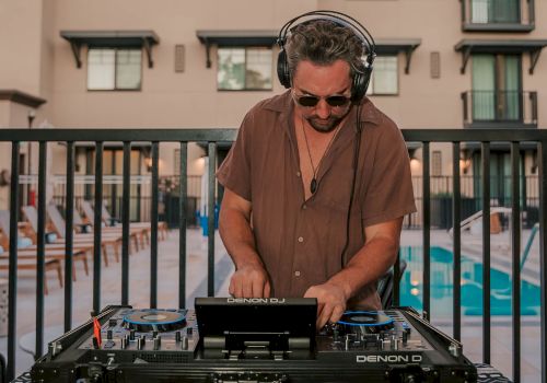A DJ adjusts controls on a mixer at an outdoor event, with colorful lights and a blurred crowd in the background.