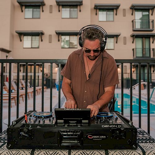 A DJ adjusts controls on a mixer at an outdoor event, with colorful lights and a blurred crowd in the background.