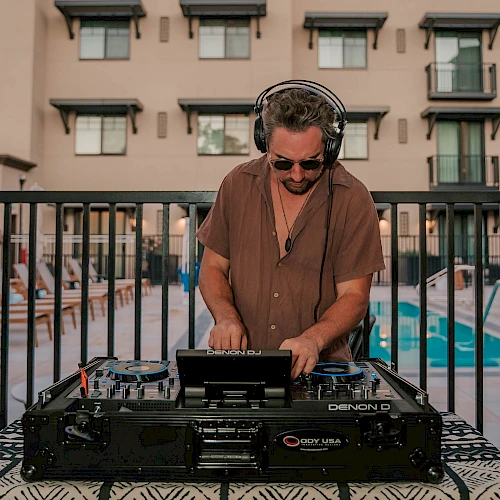 A DJ adjusts controls on a mixer at an outdoor event, with colorful lights and a blurred crowd in the background.