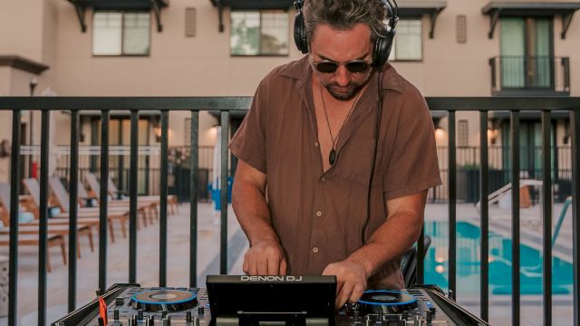 A DJ adjusts controls on a mixer at an outdoor event, with colorful lights and a blurred crowd in the background.