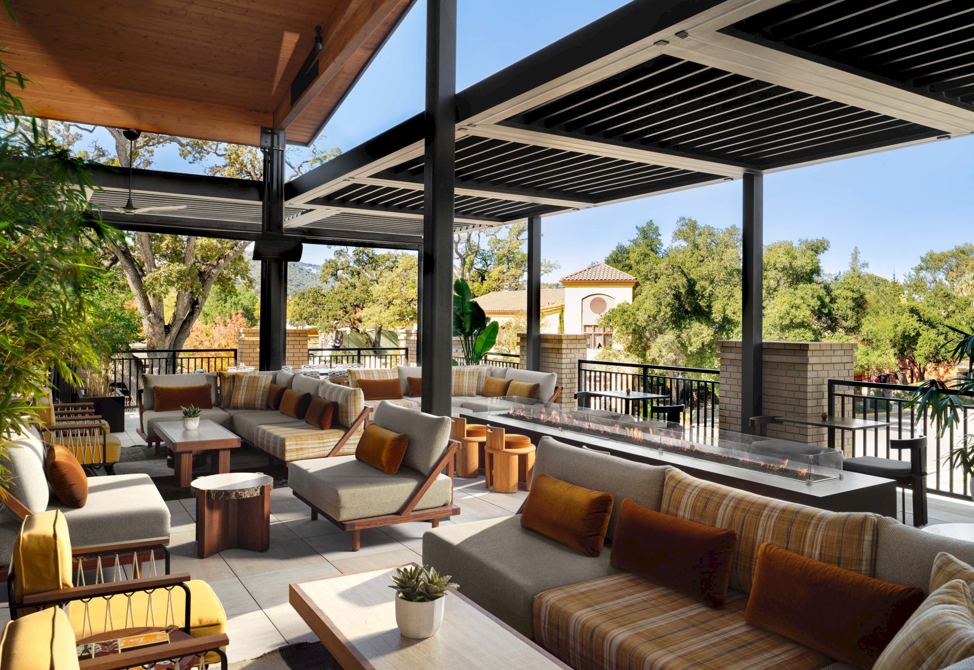 A sunny outdoor lounge with cushioned seating, wicker chairs, and small tables under a modern shaded patio, overlooking greenery.