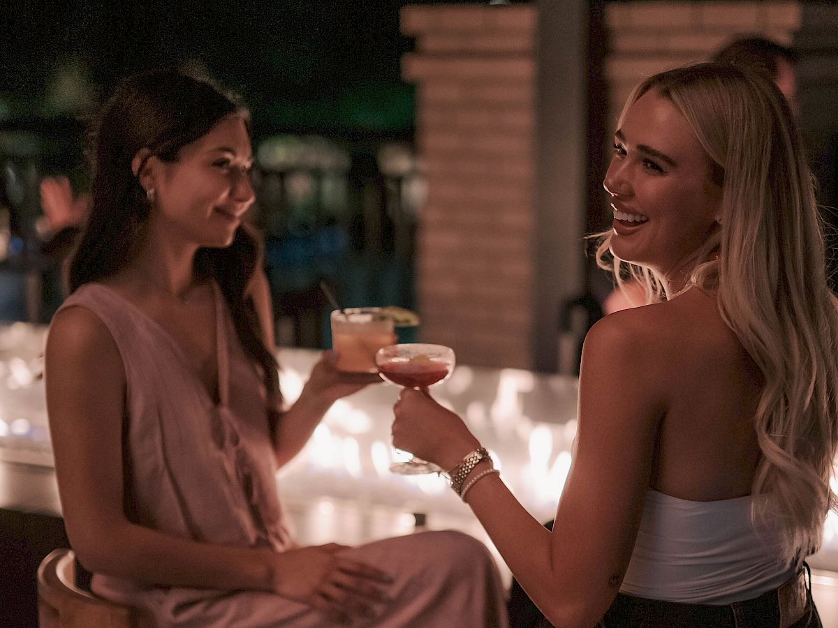 Two women sit on bar stools at night, clinking cocktails and smiling warmly at each other against soft, romantic lighting.