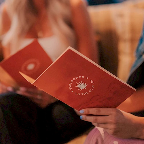 Two people sit together, holding red booklets with a white sunburst logo, likely a pamphlet or program for an event, hands visible near the bottom.
