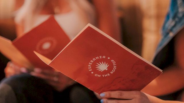 Two people sit together, holding red booklets with a white sunburst logo, likely a pamphlet or program for an event, hands visible near the bottom.