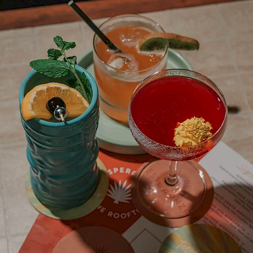 Three colorful cocktails on a table: a blue mug with a citrus slice, an orange drink with a cucumber, and a red cocktail with banana leaf garnish, all on a bright menu.