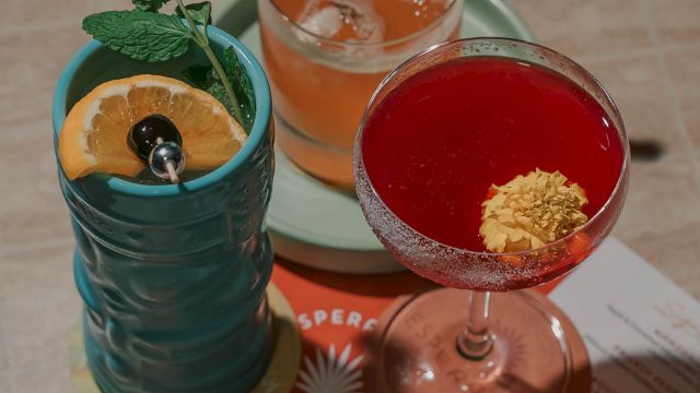 Three colorful cocktails on a table: a blue mug with a citrus slice, an orange drink with a cucumber, and a red cocktail with banana leaf garnish, all on a bright menu.