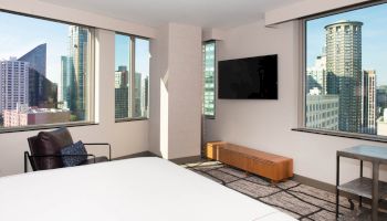 The image shows a modern hotel room with large windows, a bed, a chair, a TV on the wall, and a city skyline view outside.