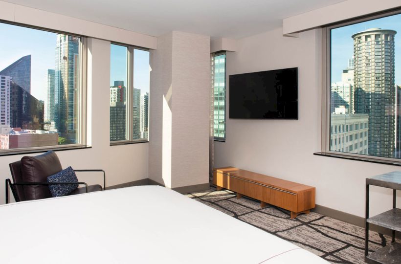 The image shows a modern hotel room with large windows, a bed, a chair, a TV on the wall, and a city skyline view outside.