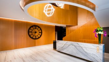 The image shows a modern reception area with a marble desk, curved wooden ceiling, wall clock, and floral arrangement.