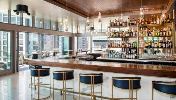 A modern, elegant bar features a long counter, stylish stools, and shelves filled with bottles against a backdrop of city views.