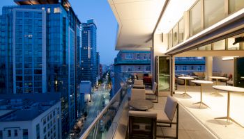 A modern rooftop terrace overlooking a city street, surrounded by tall buildings with outdoor seating and tables.