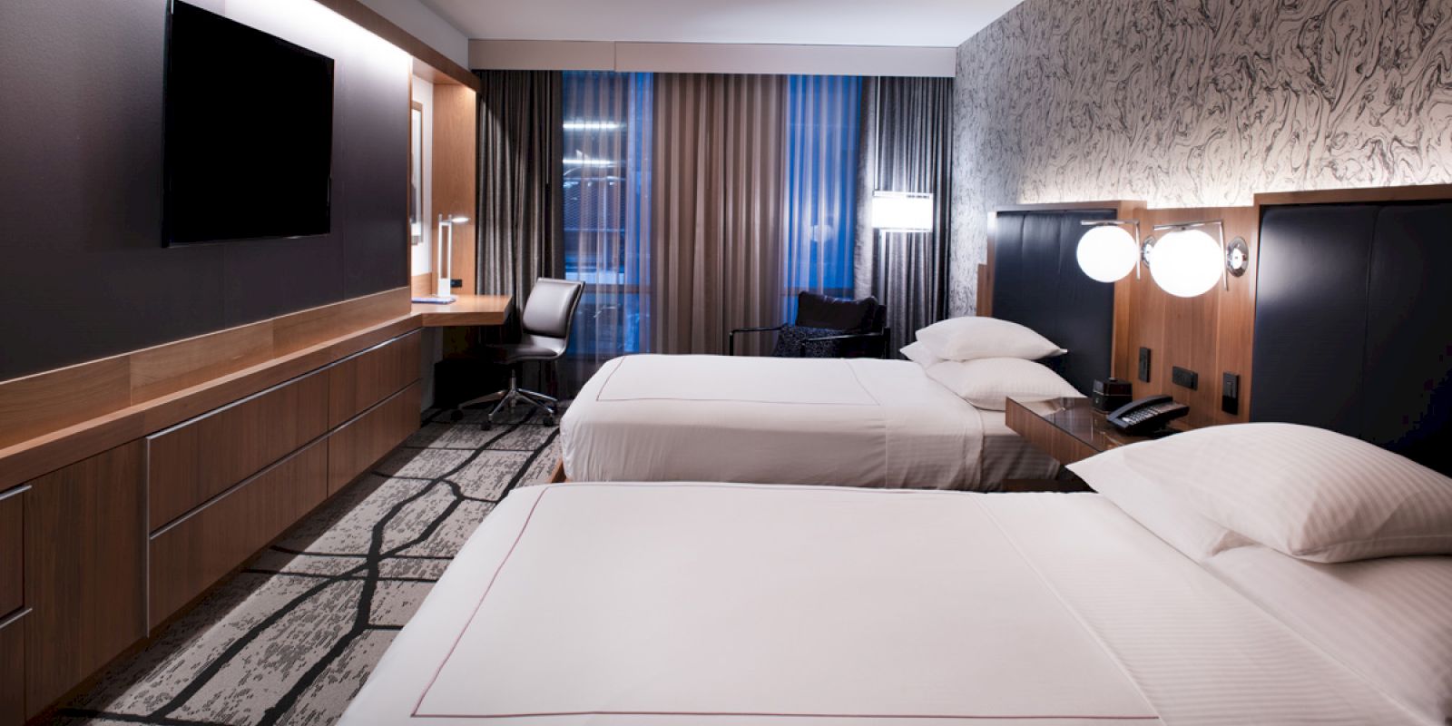 A modern hotel room with two neatly made beds, a desk, a large TV, and stylish lighting, featuring a patterned carpet and curtains.
