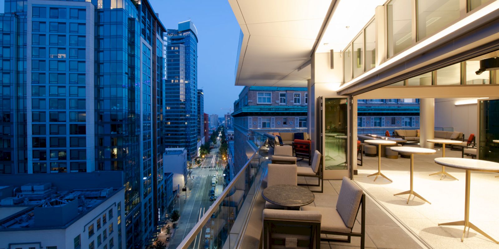 A modern rooftop terrace with seating and tables, overlooking a cityscape with tall buildings during the evening.