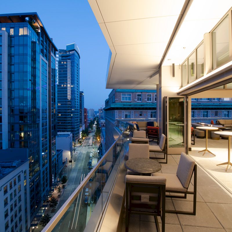 A rooftop patio at dusk with seating, overlooking city buildings and a street below.