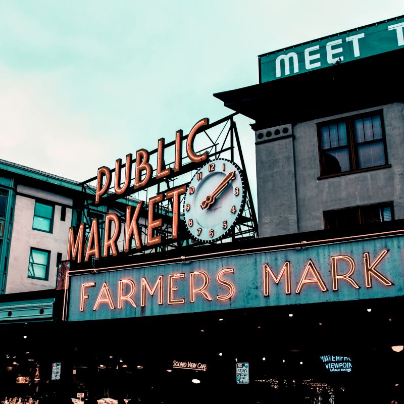 The image shows a public market with a vintage neon sign reading "Public Market" and "Farmers Market," with a clock above the text.