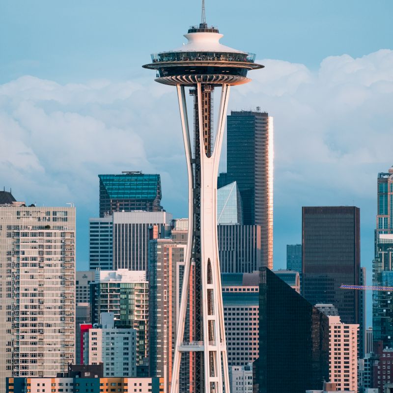 The image shows the Space Needle in front of Seattle's skyline, featuring various high-rise buildings under a blue sky.